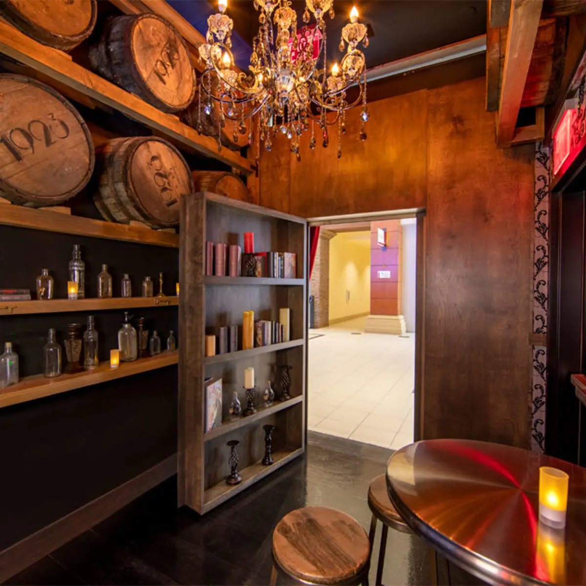 A speakeasy-style room with stacked whiskey barrels, a crystal chandelier, and a bookshelf door ajar revealing a bright hallway.