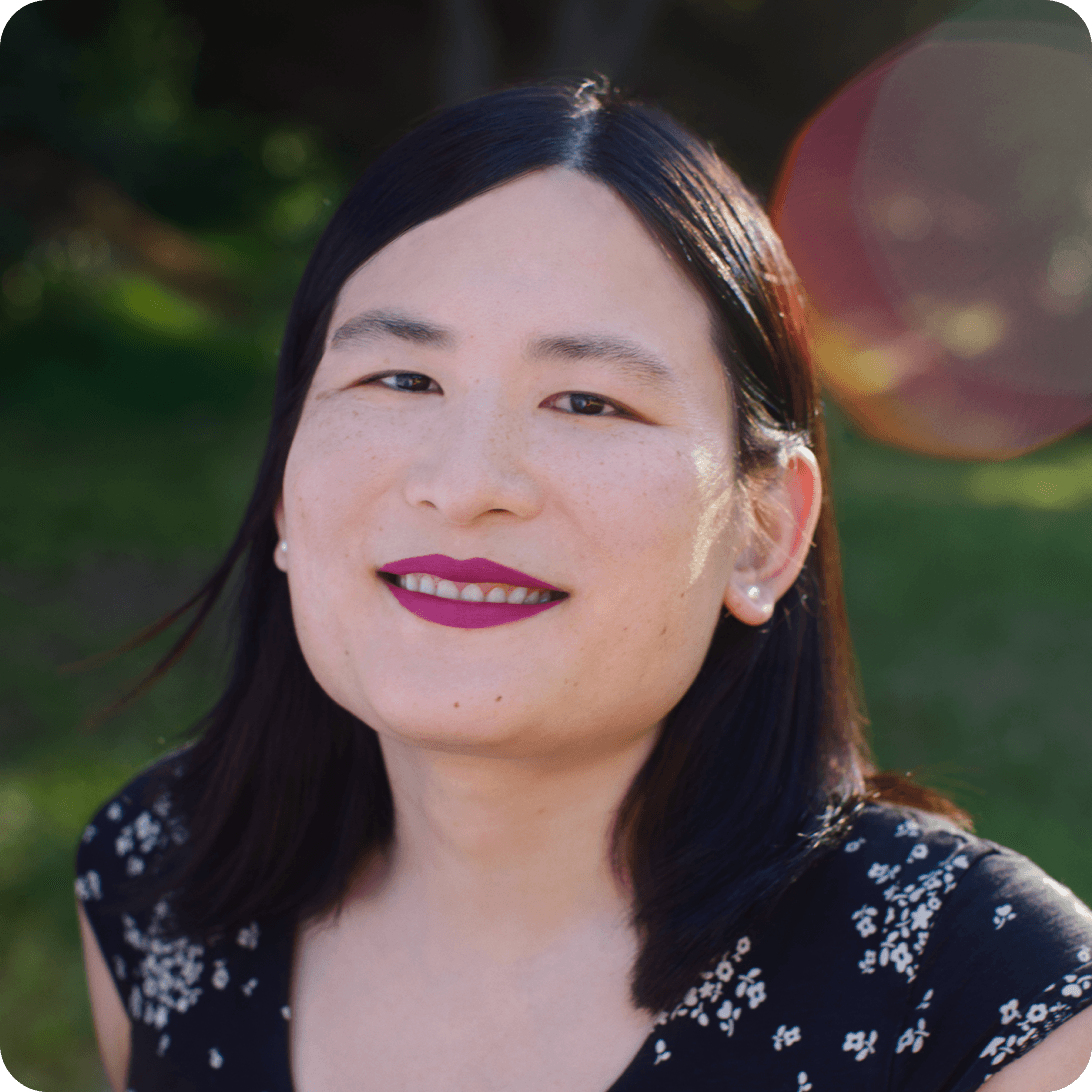 Smiling East Asian woman with dark hair, black floral top, and pearl earrings, outdoors with lens flare.