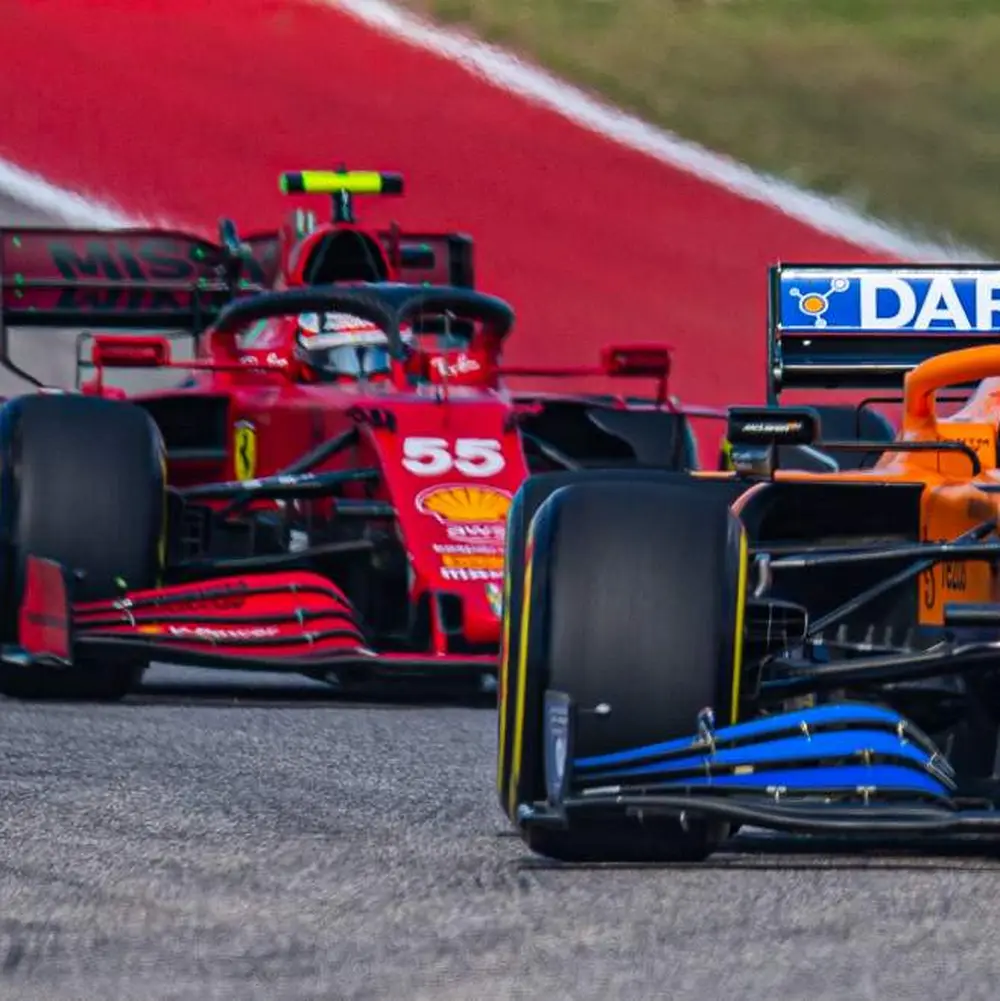 Two F1 cars racing on a track: a red Ferrari #55 behind an orange McLaren.