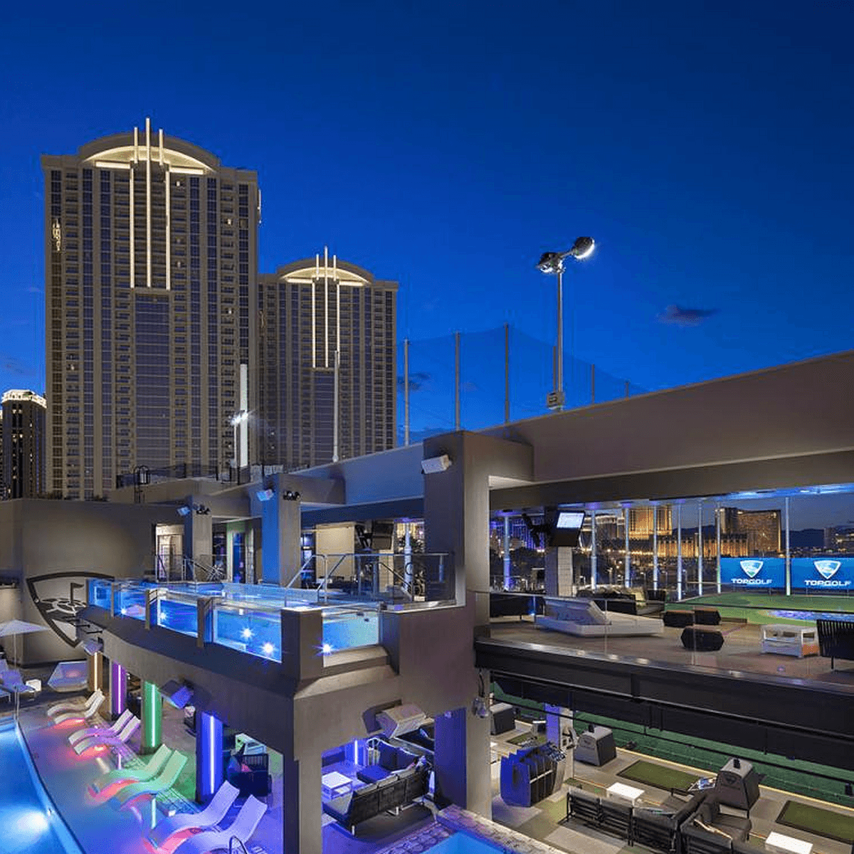 A vibrant multi-level Topgolf entertainment venue at night, featuring golf bays, illuminated pools, lounge areas, and a city skyline.